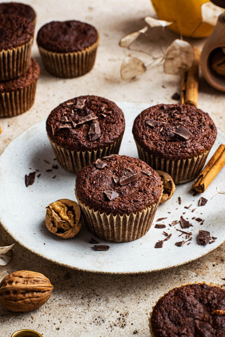 Three muffins on a plate with more around, chocolate, and nuts.