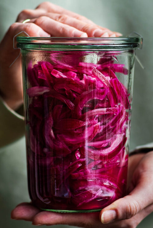 Woman holding a large jar of pickled red onions with a lid.