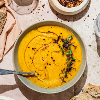 A bowl of smooth orange soup topped with olive oil and herbs, bread around.