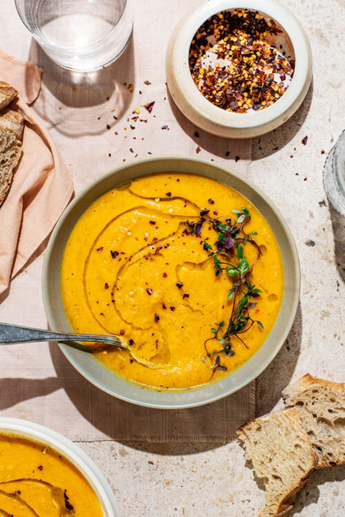 A bowl of smooth orange soup topped with olive oil and herbs, bread around.