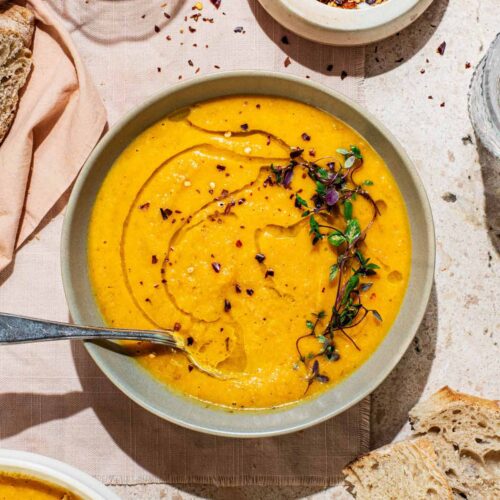 A bowl of smooth orange soup topped with olive oil and herbs, bread around.