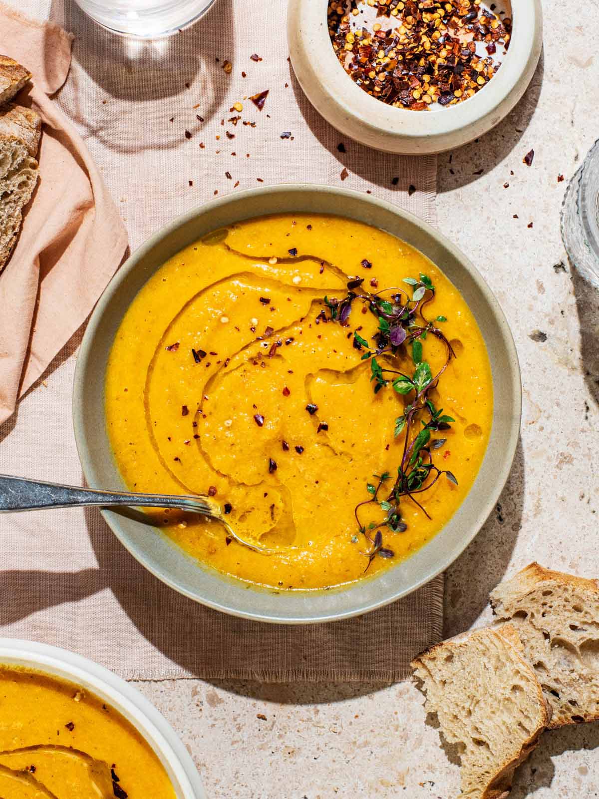 A bowl of smooth orange soup topped with olive oil and herbs, bread around.
