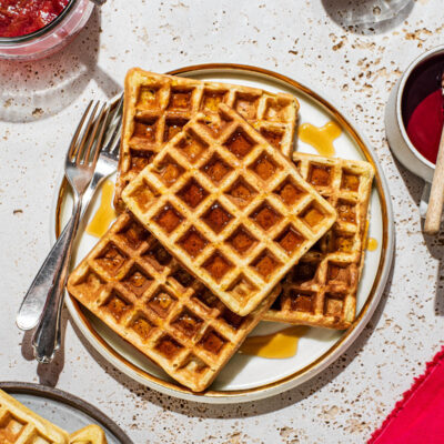 Four square waffles with maple syrup on a plate with toppings and more waffles around.