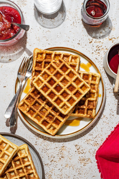 Four square waffles with maple syrup on a plate with toppings and more waffles around.