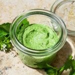 Close up of light green pesto in a glass jar with garlic mustard flowers around.