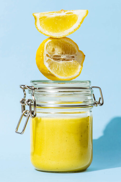 A glass jar filled with lemon vinaigrette, two lemon pieces sitting on the lid.