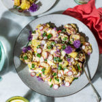 A large shallow bowl of potato salad with radishes and chive flowers, plate with more salad behind.