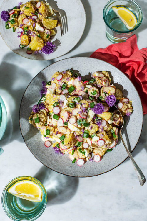 A large shallow bowl of potato salad with radishes and chive flowers, plate with more salad behind.