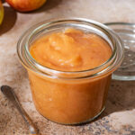 A glass canning jar filled with applesauce, apples in the background.