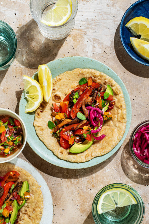 Two plates with mushroom and pepper filled tortillas, toppings in small bowls around.