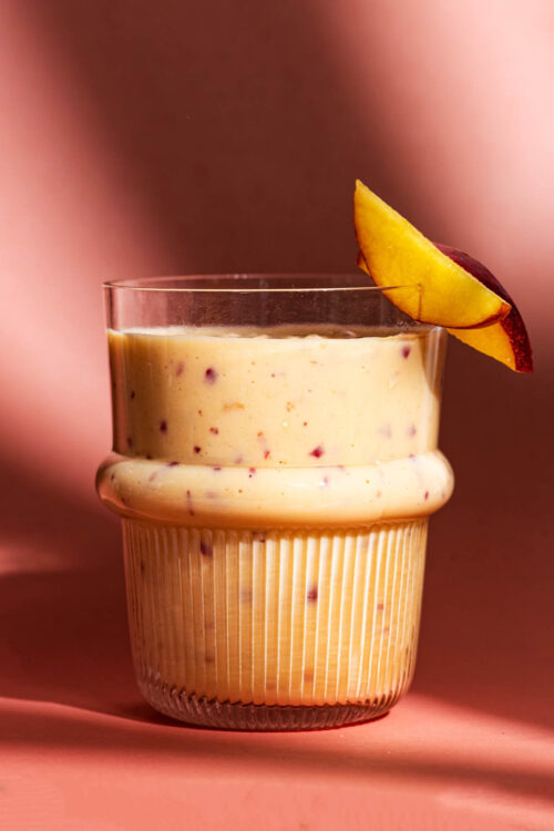 Close up of a smoothie in a glass on a pink background, with two slices of nectarine.