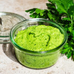 A jar of pesto with the lid off and a bunch of parsley in the background.