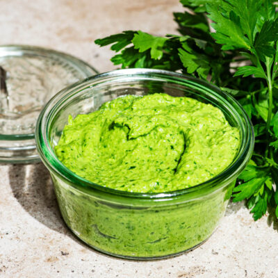 A jar of pesto with the lid off and a bunch of parsley in the background.
