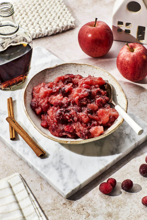 Cranberry sauce in a bowl with apples, maple syrup, and cinnamon around.