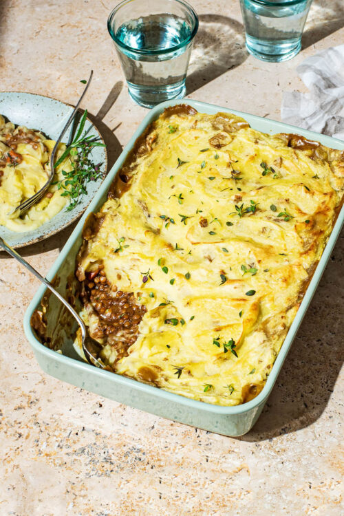 A lentil cottage pie in a rectangular baking dish, with one scoop removed to see the filling under the mashed potatoes.