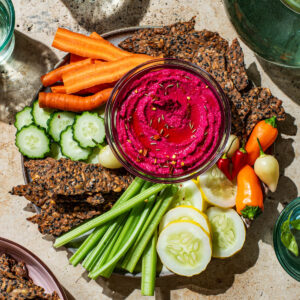 A serving plate with beet hummus and a selection of raw vegetables and seed crackers.