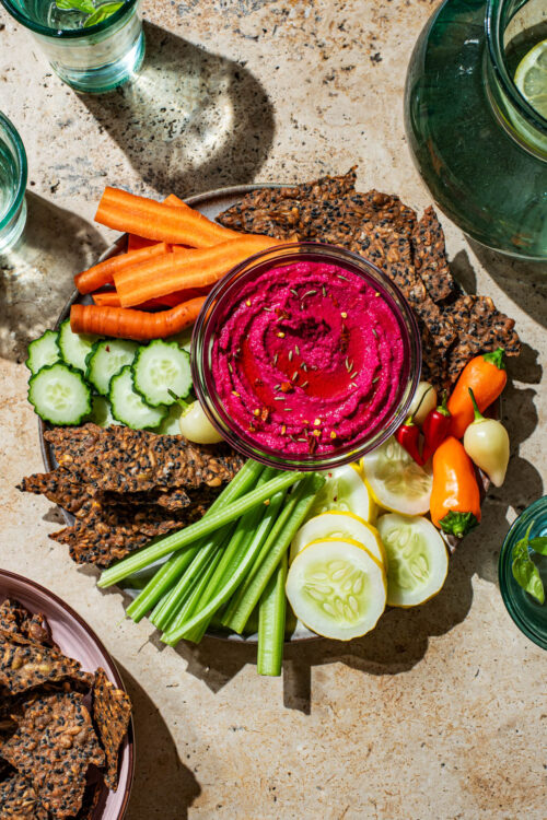 A serving plate with beet hummus and a selection of raw vegetables and seed crackers.