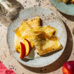 Two flat pancakes folded into triangles on a plate with fruit and yogurt.