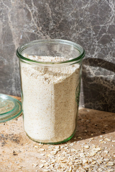 A glass jar full of oat flour, with rolled oats around.