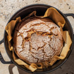 A round loaf of bread with a crackled top in a parchment-paper lined Dutch oven pot.