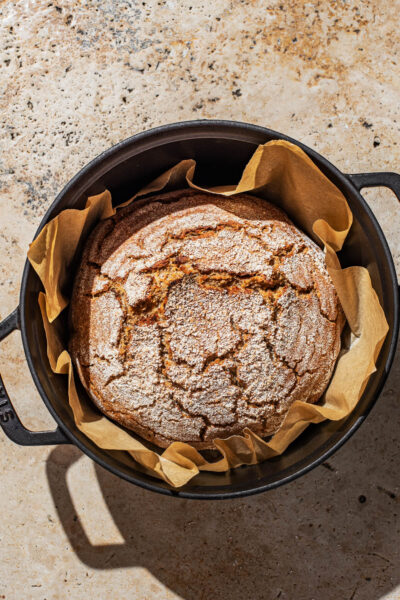 A round loaf of bread with a crackled top in a parchment-paper lined Dutch oven pot.