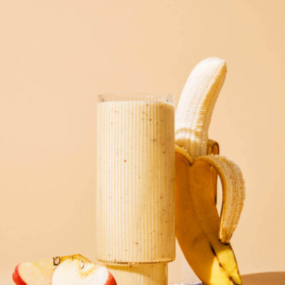 A peach-coloured smoothie in a glass with cut apple and half-peeled banana beside.