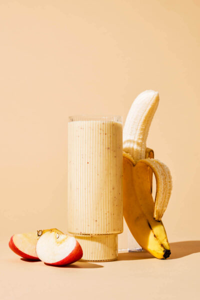 A peach-coloured smoothie in a glass with cut apple and half-peeled banana beside.