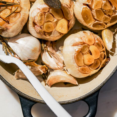 Several heads and cloves of roasted garlic in an oven dish with herbs.