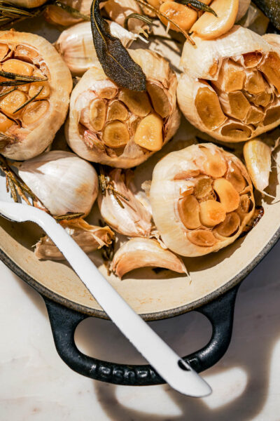 Several heads and cloves of roasted garlic in an oven dish with herbs.