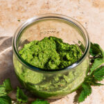 A jar of pesto with the lid off, stinging nettle leaves beside the jar.