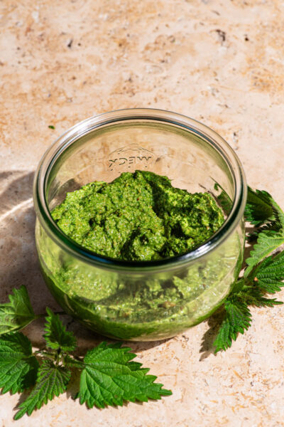 A jar of pesto with the lid off, stinging nettle leaves beside the jar.