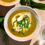 Top-down view of a green-brown soup in bowls with cream and herbs.