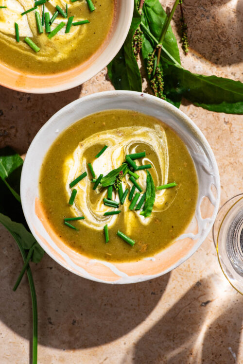Top-down view of a green-brown soup in bowls with cream and herbs.