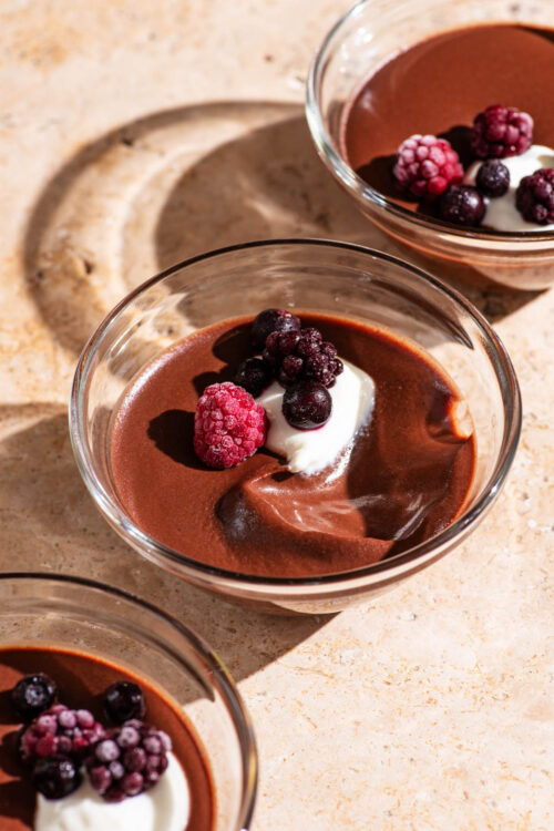 Three small glass bowls filled with chocolate pudding, garnished with cream and berries.