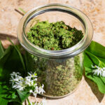 A jar full of green salt with wild garlic leaves and flowers beside.