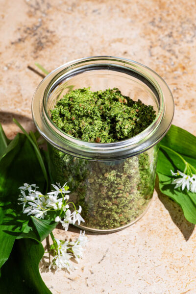 A jar full of green salt with wild garlic leaves and flowers beside.