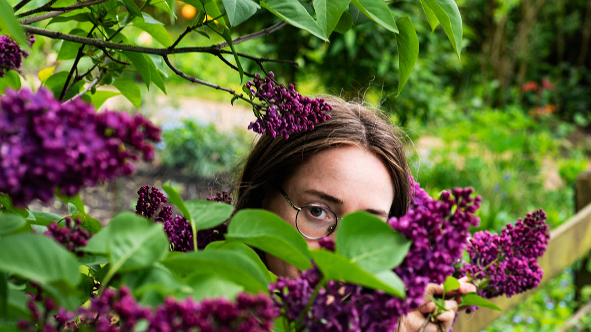 The head of a bespectacled woman half-hidden behind a lilac bush.