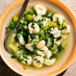 A bowl of soup with tortellini, peas, beans, and spinach.