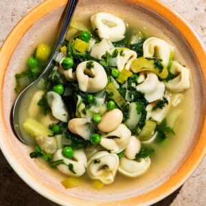 A bowl of soup with tortellini, peas, beans, and spinach.