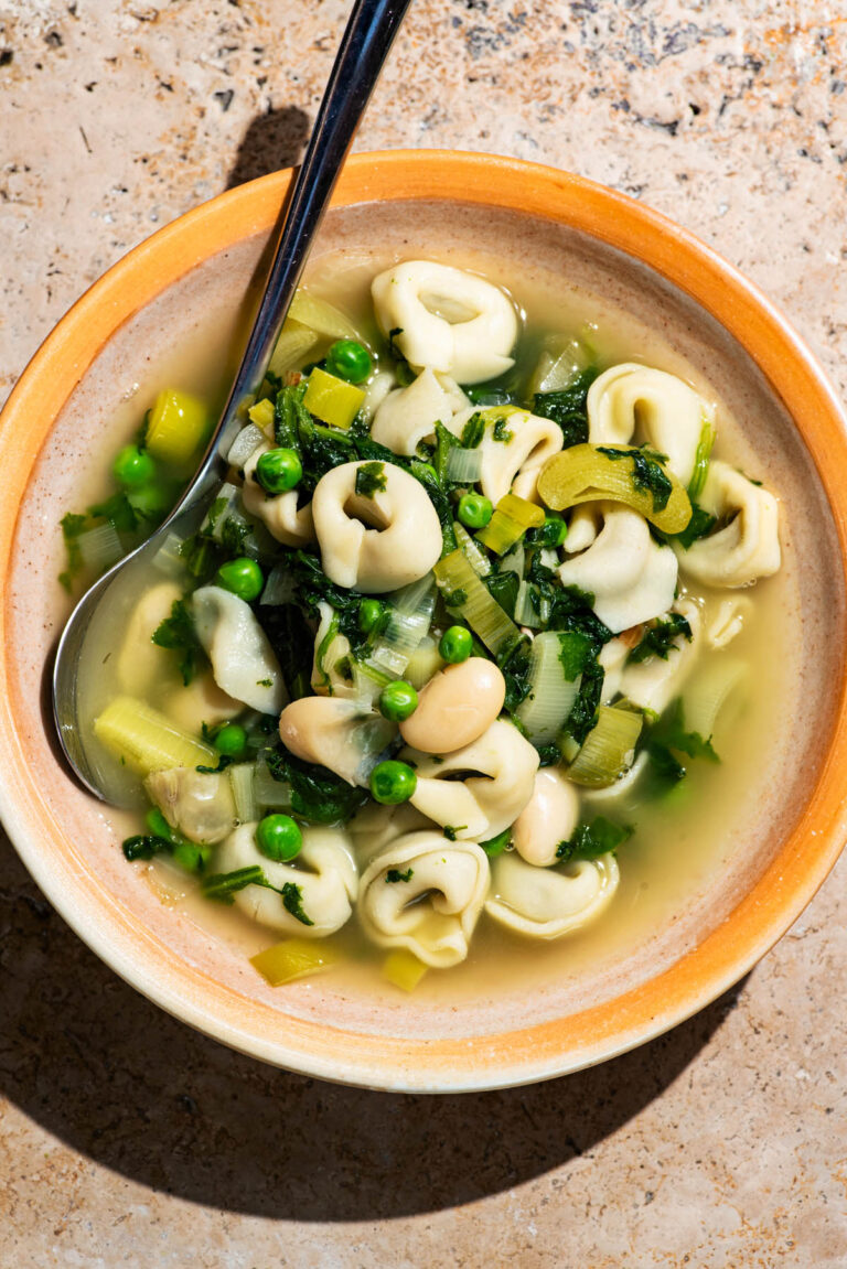 A bowl of soup with tortellini, peas, beans, and spinach.