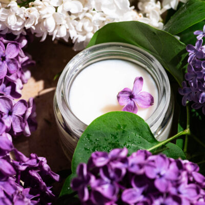 A small jar of white cream surrounded by lilac blossoms.