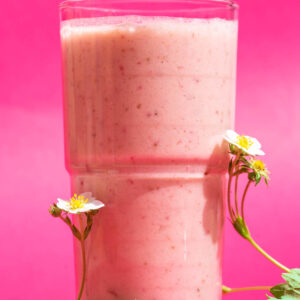 Close-up of a pink smoothie in a clear glass with strawberry flowers beside.