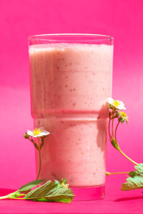 Close-up of a pink smoothie in a clear glass with strawberry flowers beside.
