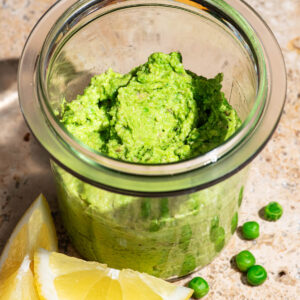 A light green spread in a glass jar, with lemon and peas in front.