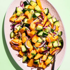 A chopped salad with orange melon, cucumber, and red onion on a pink platter.