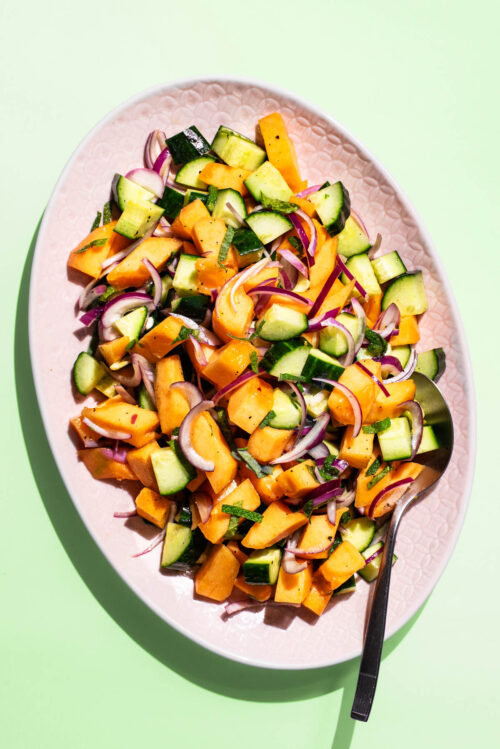 A chopped salad with orange melon, cucumber, and red onion on a pink platter.