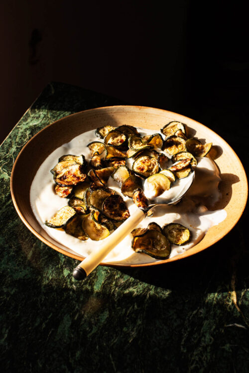 A plate of golden zucchini slices on yogurt in hard sunlight.