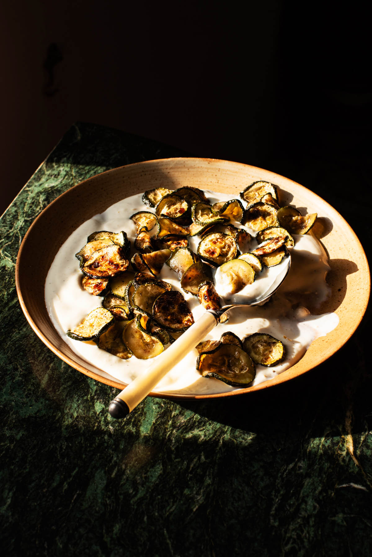 A plate of golden zucchini slices on yogurt in hard sunlight.