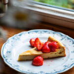 A slice of dairy-free cheesecake topped with strawberries on a plate on a windowsill.