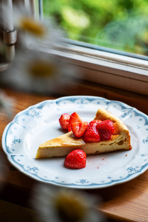 A slice of dairy-free cheesecake topped with strawberries on a plate on a windowsill.